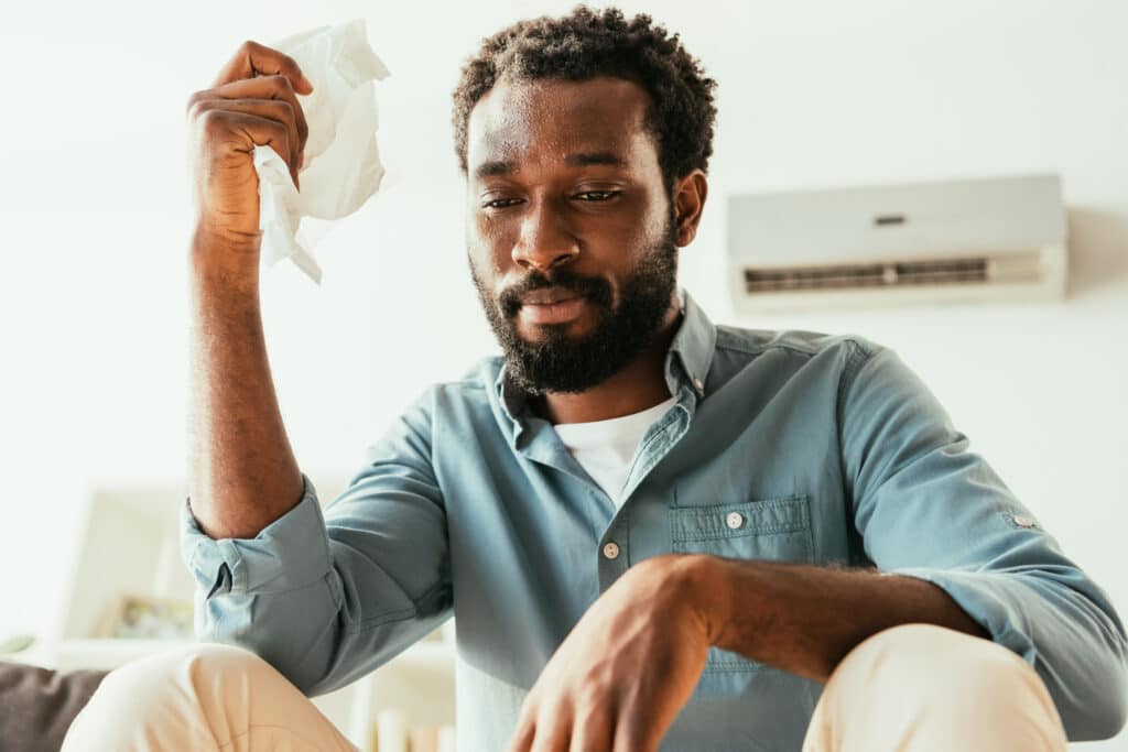 unhappy african american man holding napkin while suffering from summer heat at home Mann vor Klimaanlage